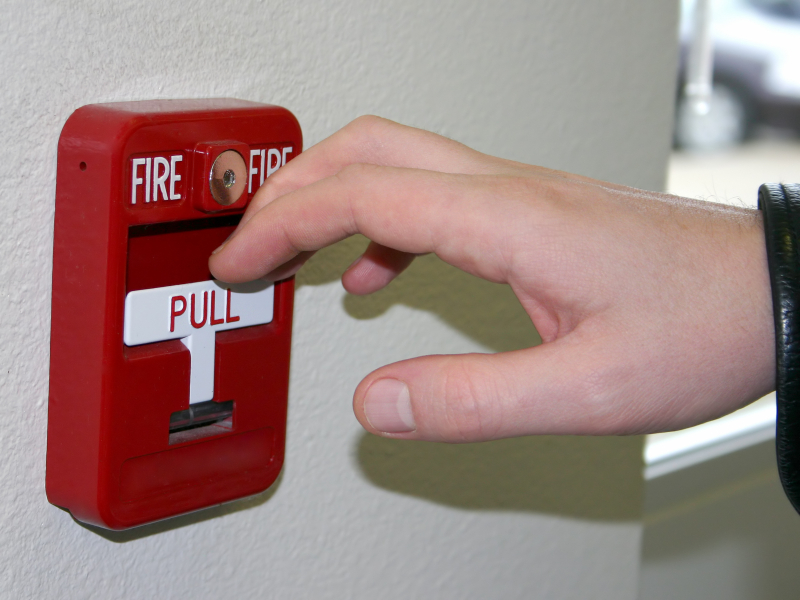 A hand reaches to pull the tab on a fire alarm.